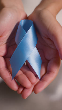 Vertical-Video-Close-Up-Of-Person-Holding-Blue-Ribbon-Symbolizing-Awareness-Of-Men's-Health-And-Cancer